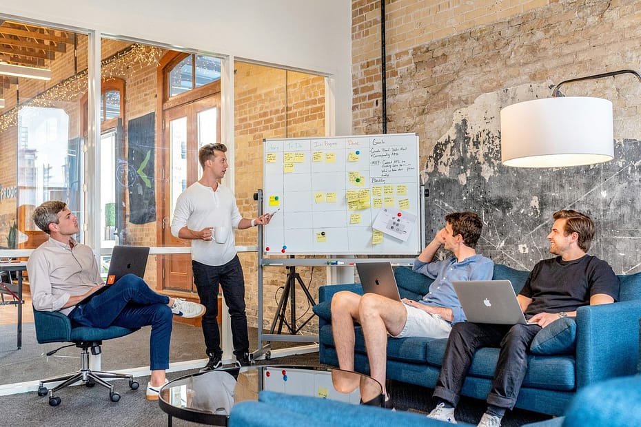 three men sitting while using laptops and watching man beside whiteboard